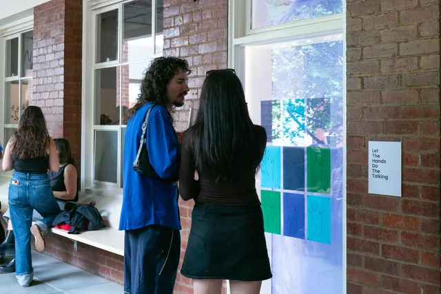 Two people standing in front of a white window displaying various sheets of coloured A4 paper with hands drawn on them.