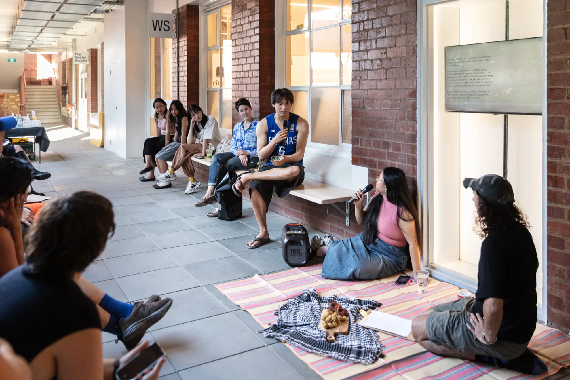 There is a crowd of people seated on benches that line an open walkway, built of brick and large white windows. One figure on the bench is holding a microphone and gesturing toward another microphone holder sitting on a rub near them. The rug is shared with another figure. Everyone is listening intensely to the elevated figure with the microphone in dialogue.