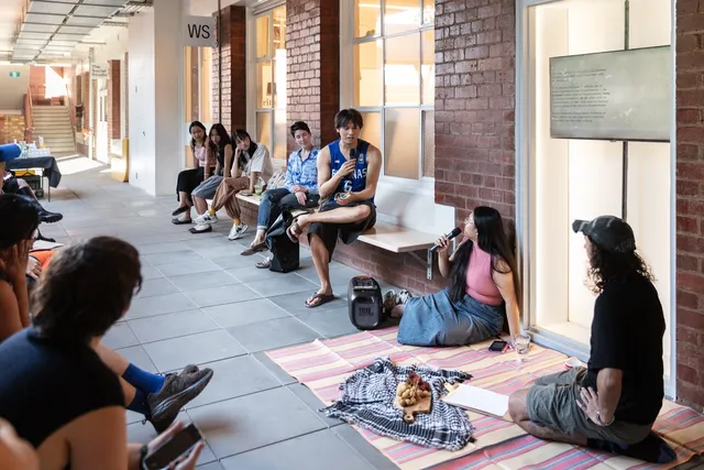 There is a crowd of people seated on benches that line an open walkway, built of brick and large white windows. One figure on the bench is holding a microphone and gesturing toward another microphone holder sitting on a rub near them. The rug is shared with another figure. Everyone is listening intensely to the elevated figure with the microphone in dialogue.