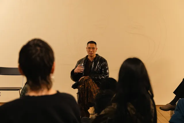 An individual sits on a chair on stage, talking to the audience. There is a chair to the left of him. We see the figures of two audience members from behind who are listening.