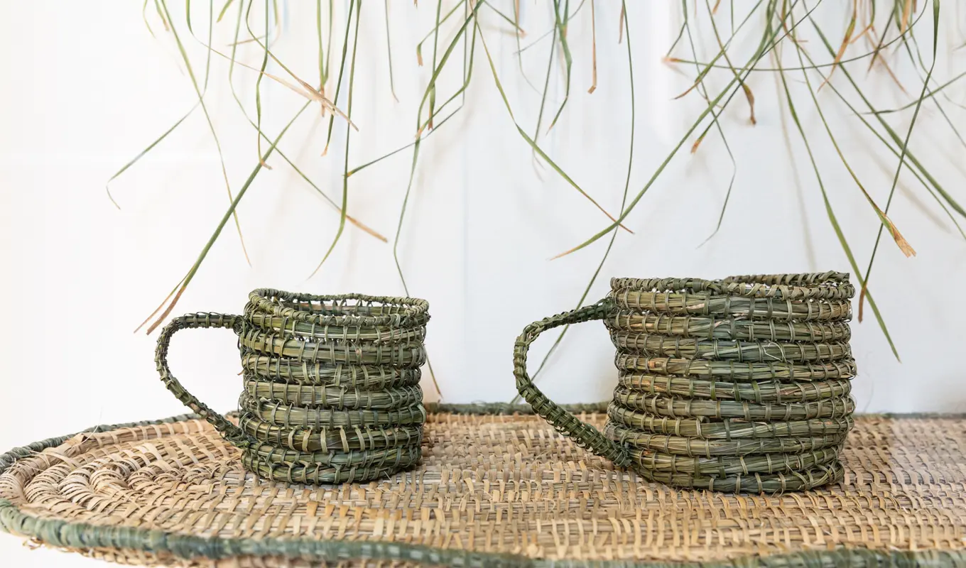 Some densely woven mugs in colours of green, tinted yellow and brown, natural fibres on a rounded, weaved platform. There are 2 mugs, only half of the overall composition install. Hung above the elevated mugs is fragments, is the fibre in it's natural state - draping.