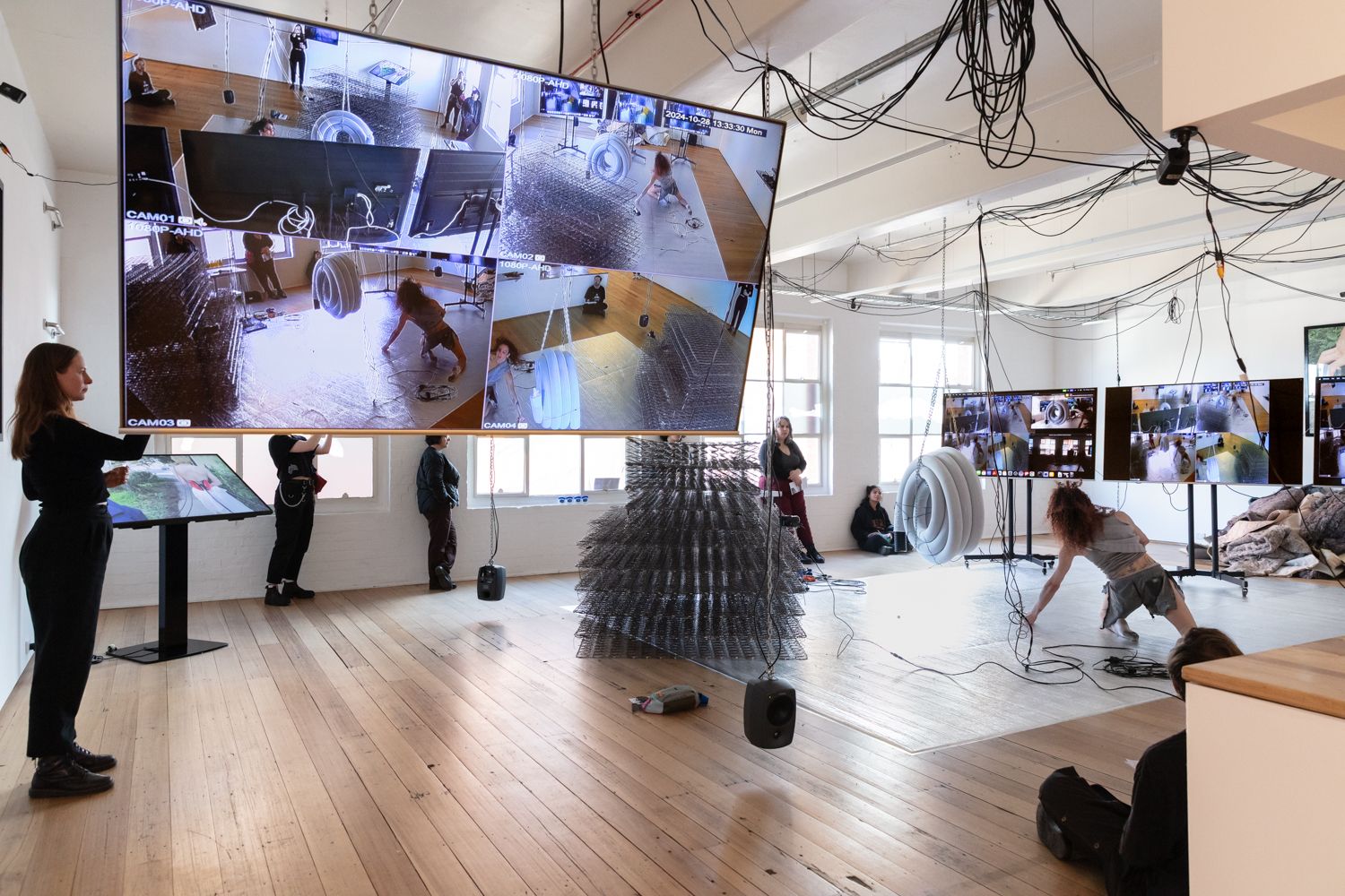 A woman is performing in the middle of the gallery. Visitors are watching against the walls. The performance is also displayed on the four screens in the gallery.