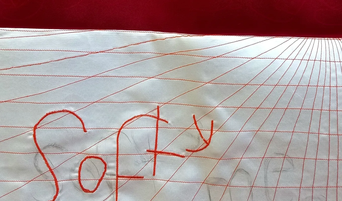 The top half of the image is a deep red and the bottom half features an embroidered grid that distorts into the background. The word 'softy' is embroidered on top of the grid with faint pencil markings of the words 'softy' and 'men'.