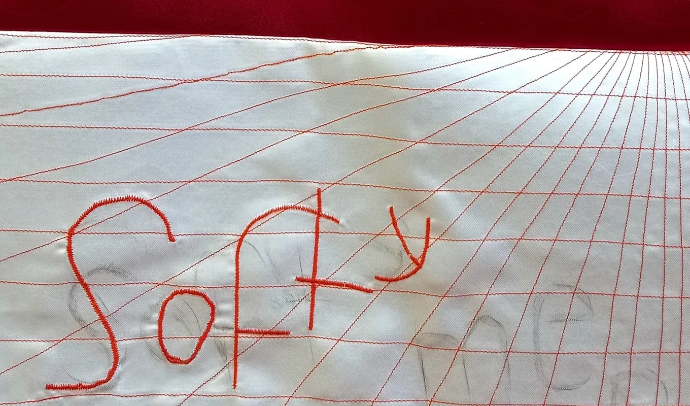The top half of the image is a deep red and the bottom half features an embroidered grid that distorts into the background. The word 'softy' is embroidered on top of the grid with faint pencil markings of the words 'softy' and 'men'.