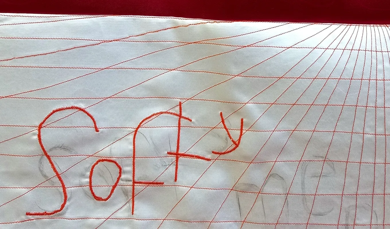 The top half of the image is a deep red and the bottom half features an embroidered grid that distorts into the background. The word 'softy' is embroidered on top of the grid with faint pencil markings of the words 'softy' and 'men'.