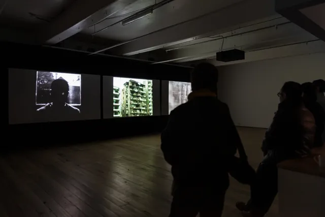 Gallery visitors stand in a dark gallery observing a three channel video work being projected in the gallery by the artist.