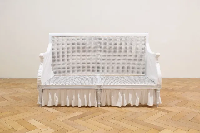 Front view of Fiona Abicare's white couch sculpture. The couch frame is provincial in style and made from wood that has been painted white. The arms have been covered in white fabric and at the bottom of the couch is a frill of white gathered fabric.