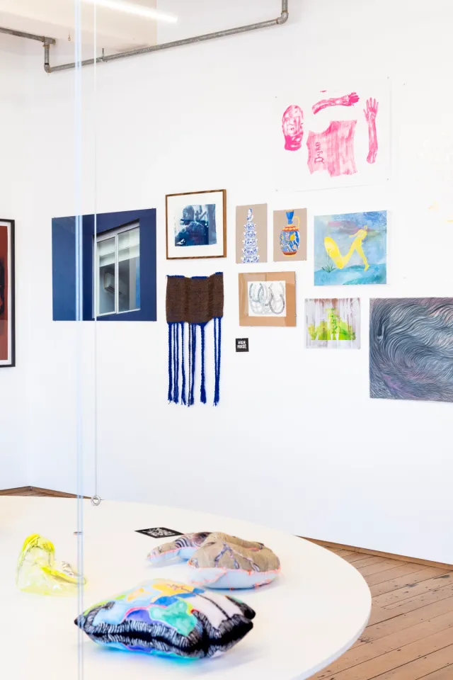 A collection of various works are assembled in a salon hang on the wall of West Space as part of the exhibition "Kin". There is a floating white circular shelf in the front of the image with some soft sculptures on it.