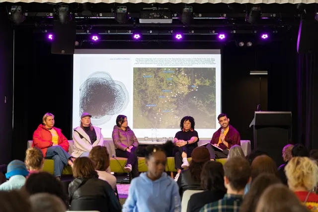 A panel of people sit on a stage in front of an illuminated projection screen, they are facing the audience and we can see the back of the audiences heads
