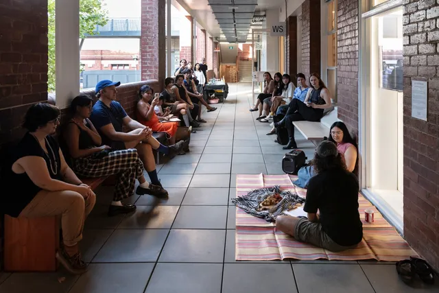 two long rows of people sit at the edges of an open corridor, on both sides. There is a carpet in the foreground where tow people are talking. Everyone's gases are directed toward these figures in discussion, who in front of them lies a blanket-like cloth full of plant matter or materials. The even is taking place out the front of West Space.