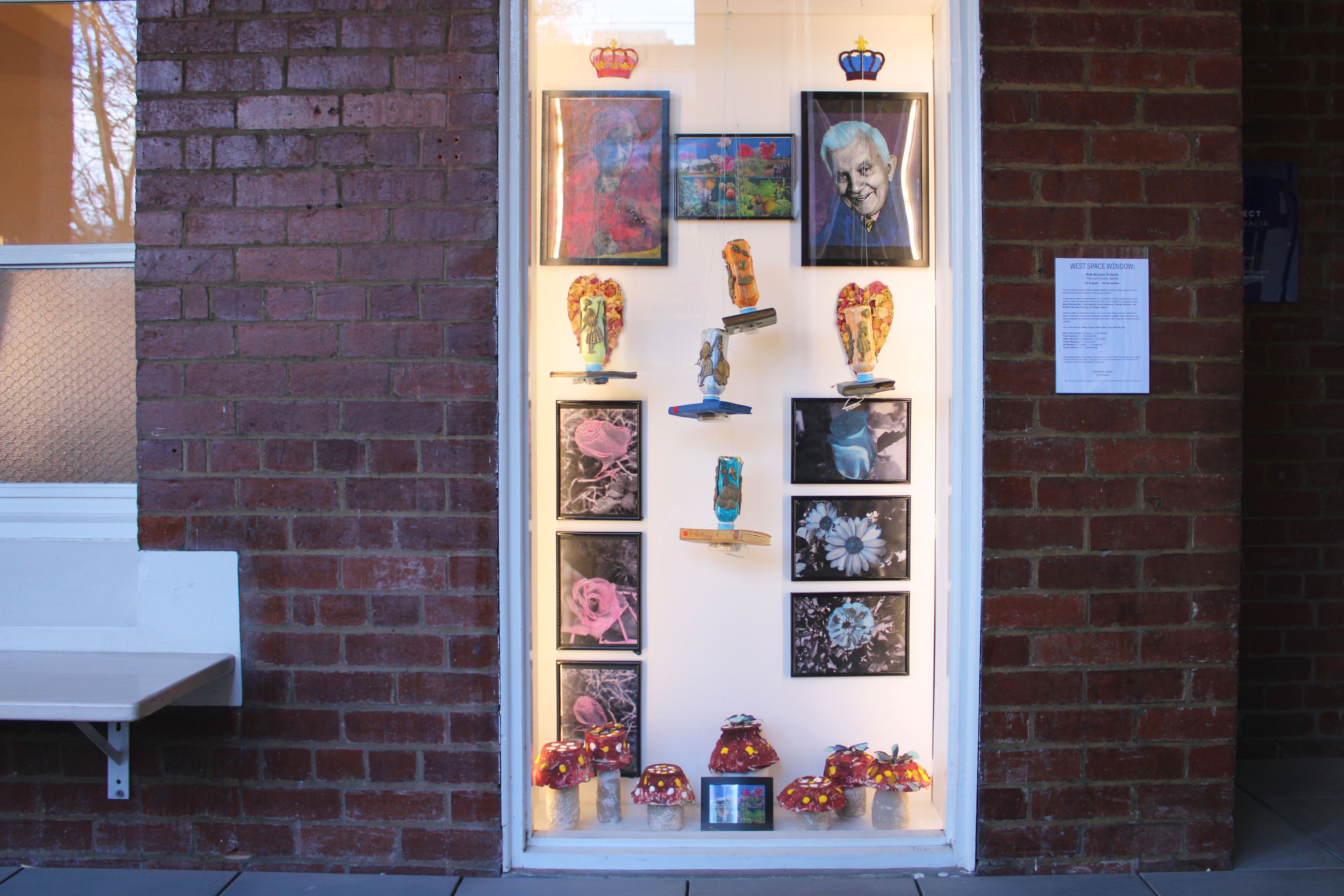 Behind a white framed window there are a series of 10 black framed colour manipulated photographic images, amongst them there are various multicoloured sculptural works scattered. Above the figures are one red and one blue crown.