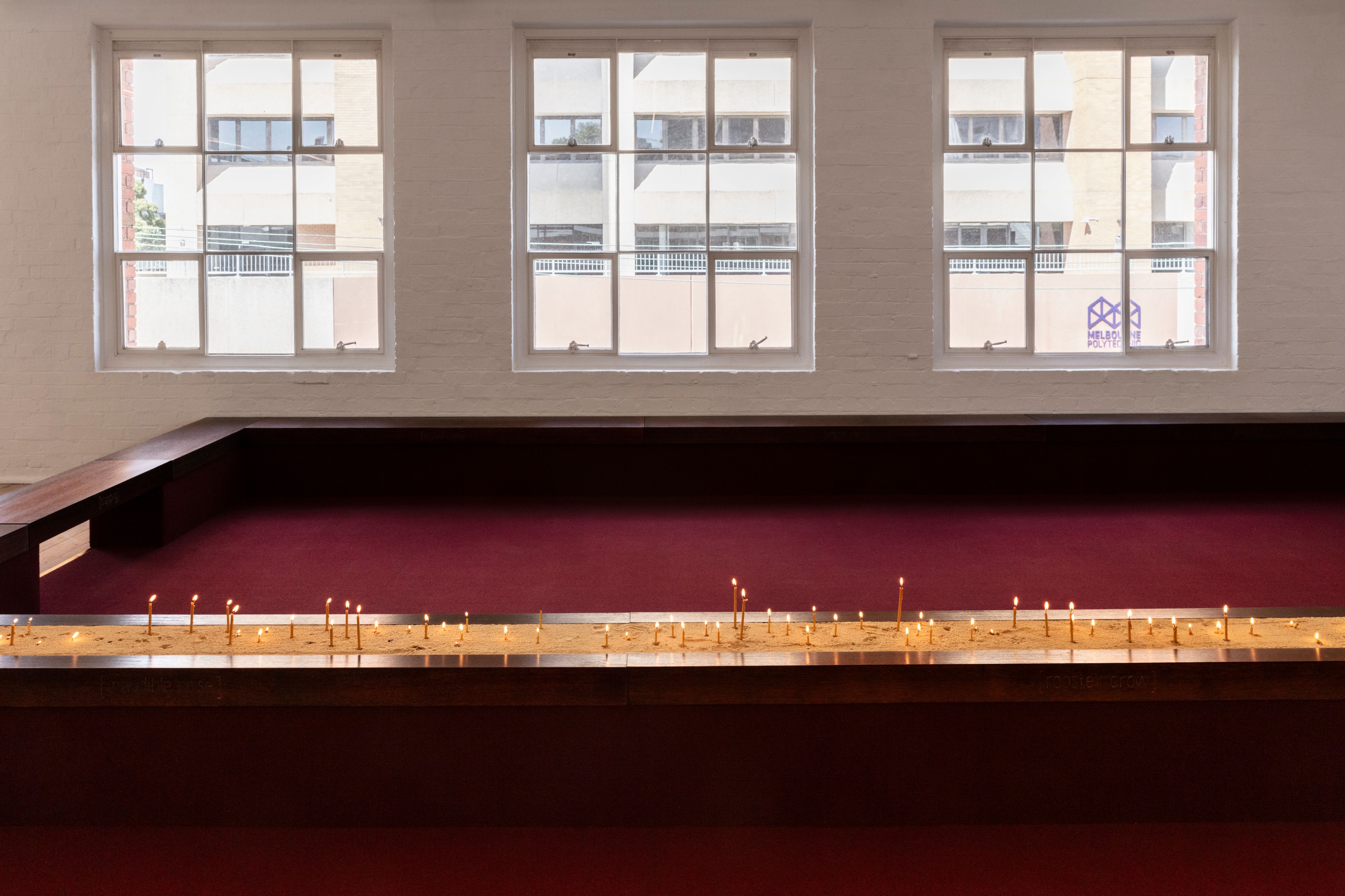 Long wooden bench structure with sand and lit candles sits in front of three large nine grid windows.