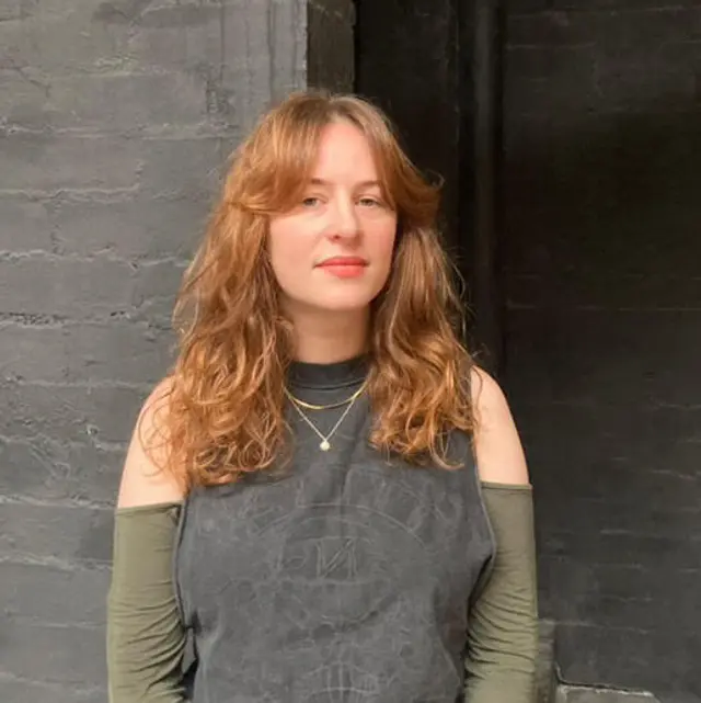 A head shot of a person with long wavy reddish hair and a grey top with olive green sleeves