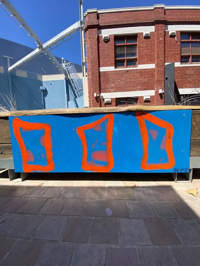 A rectangular board painted bright blue with three bright orange abstract geometric forms painted on top.