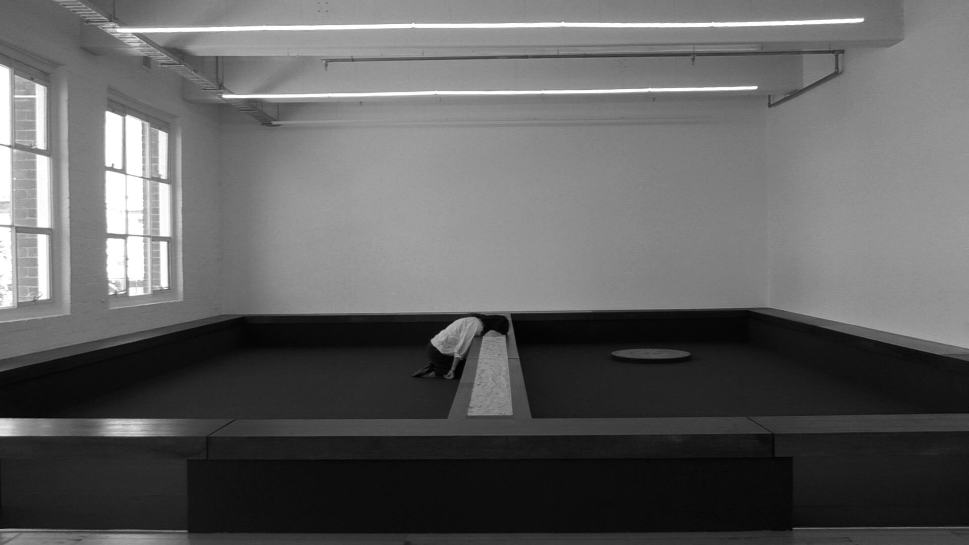 Black and white image of Tina Stefanou kneeling on the ground with her head buried in sand in the centre of her wooden installation for Motet Fail.