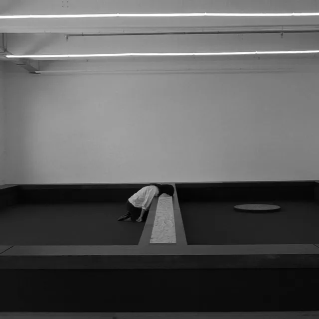 Black and white image of Tina Stefanou kneeling on the ground with her head buried in sand in the centre of her wooden installation for Motet Fail.