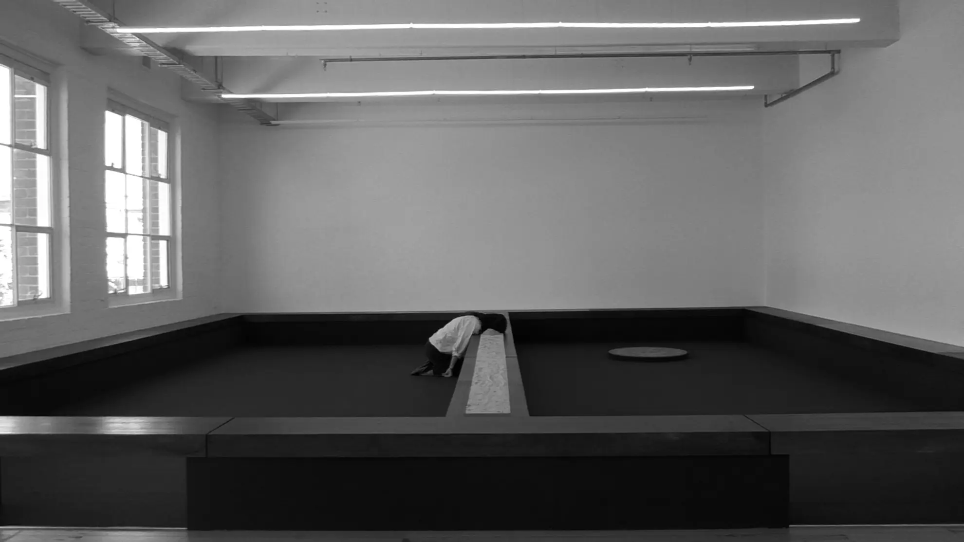 Black and white image of Tina Stefanou kneeling on the ground with her head buried in sand in the centre of her wooden installation for Motet Fail.