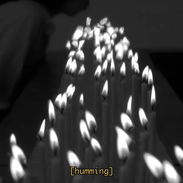 a black and white film still on a long bed of burning candles. In the background there is a shadowed figure on her knees leaning forward to blow on the candles. A subtitle reads "humming"