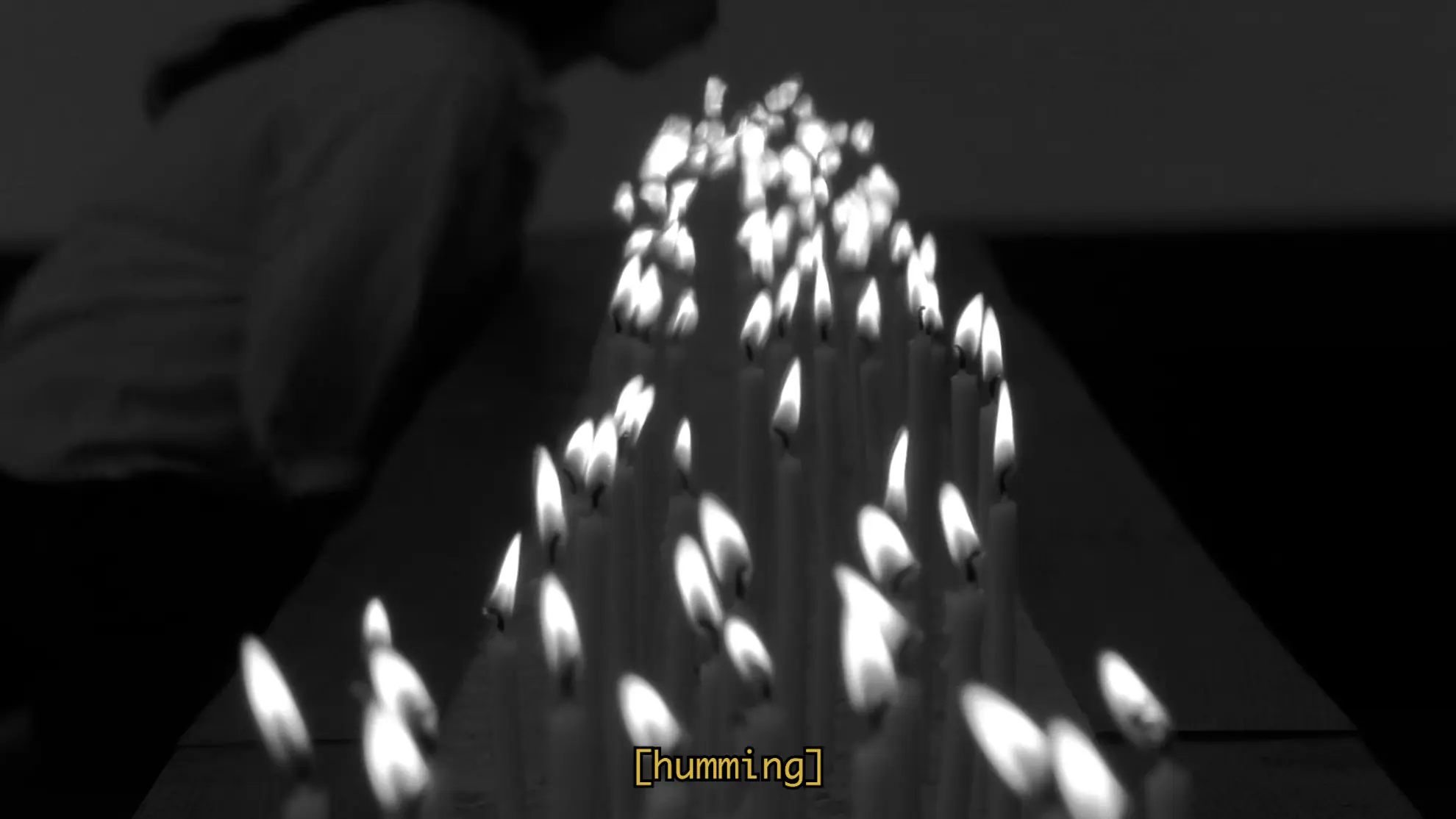 a black and white film still on a long bed of burning candles. In the background there is a shadowed figure on her knees leaning forward to blow on the candles. A subtitle reads "humming"