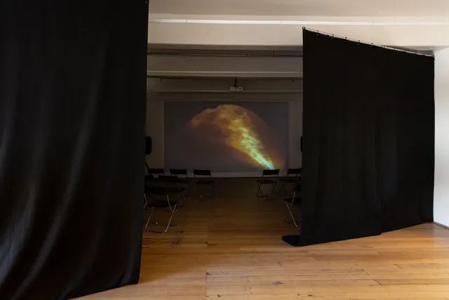 A projected image is shown through two black curtains hanging in the gallery. The film still shows a blurred curved image of trees and fire casting an amber light through darkness.