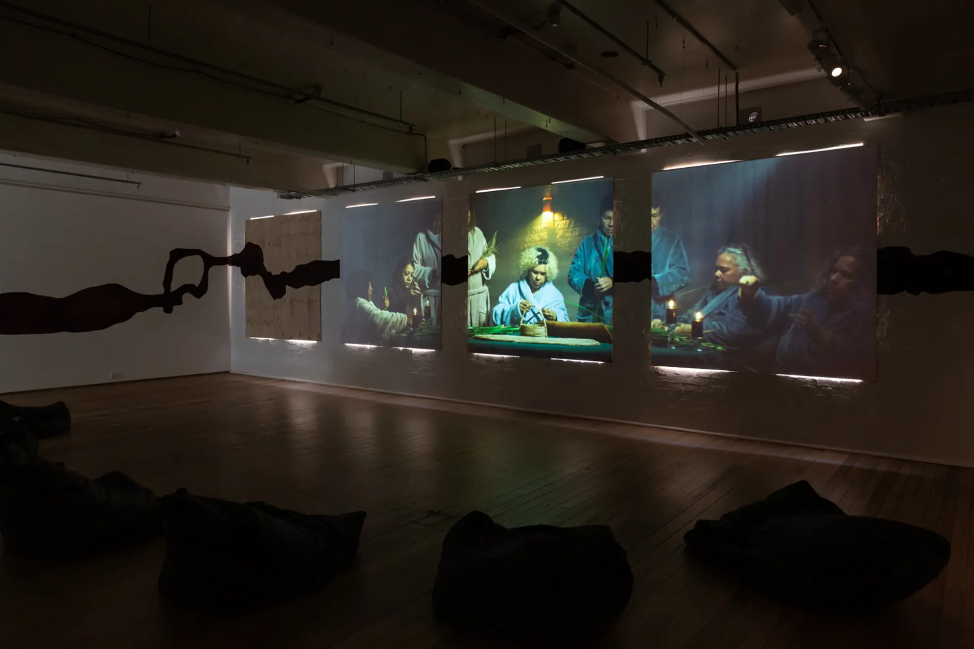 Three large projection screens displaying scenes of people sitting around a table weaving, with a shadowy black silhouette running horizontally across the wall. The room has wooden flooring and several black bean bags positioned in front of the projections.