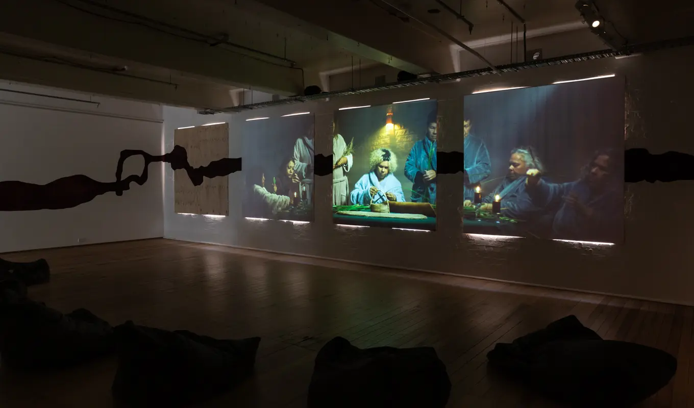 Three large projection screens displaying scenes of people sitting around a table weaving, with a shadowy black silhouette running horizontally across the wall. The room has wooden flooring and several black bean bags positioned in front of the projections.