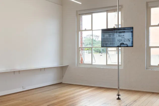 The image is of a screen suspended at eye level in the West Space Gallery showing Guled Abdulwasi's "The Block" (2019). To the left is part of a shelf suspended off of the wall with a series of photographs placed on it.