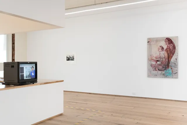 Installation view of West Space. On the left side of the image, a CRT television rests on a white counter and displays a person in a blue jumper standing in front of a portrait. The middle of the photo shows a small black and white image mounted on a white wall. On the right of the image is a large painting depicting a figure seated at a table with another standing behind them.