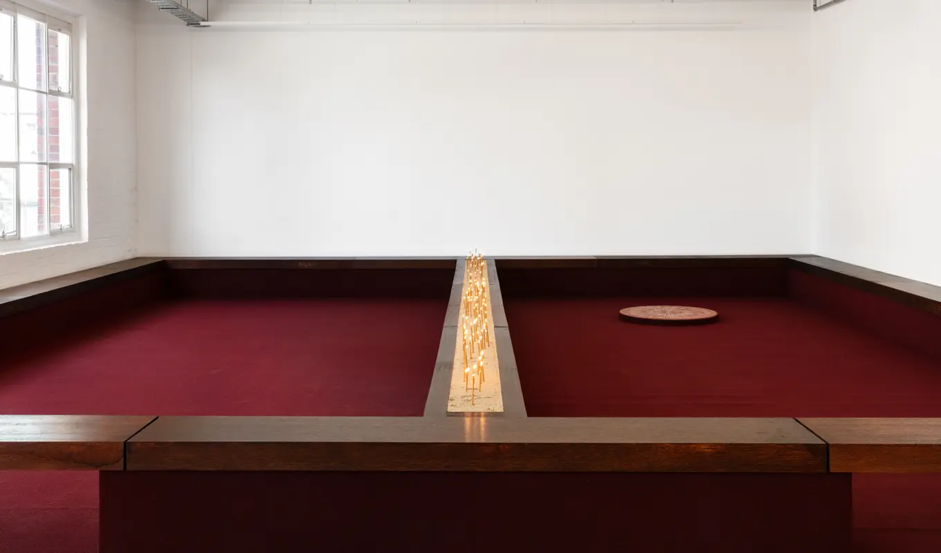Installation view of Motet Fail featuring a wall to wall seating structure with a divided ledge down the middle containing a bed of sand filled with candles. The whole structure is lined with burgundy carpet. On the right there is a large circular disc on the carpet.