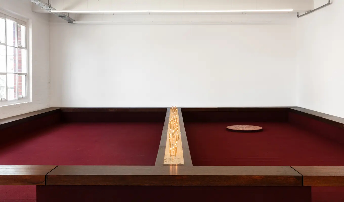 Installation view of Motet Fail featuring a wall to wall seating structure with a divided ledge down the middle containing a bed of sand filled with candles. The whole structure is lined with burgundy carpet. On the right there is a large circular disc on the carpet.