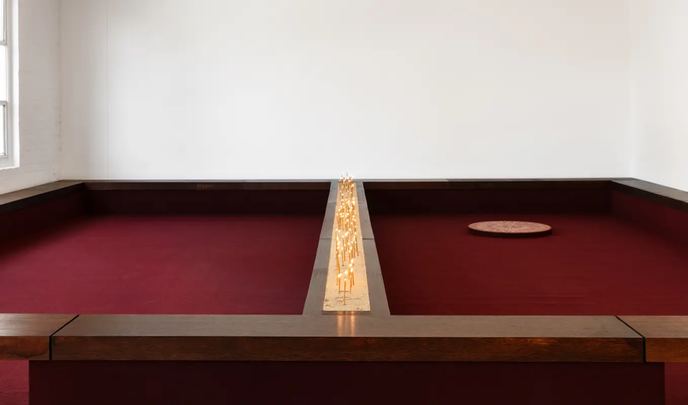 Installation view of Motet Fail featuring a wall to wall seating structure with a divided ledge down the middle containing a bed of sand filled with candles. The whole structure is lined with burgundy carpet. On the right there is a large circular disc on the carpet.