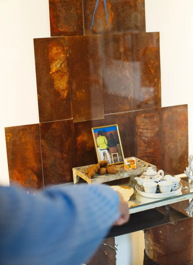 A small teacup set on a ceramic tray, two cigarettes on a flat shell, a small box with some orange graphics on it, some small kiwi-fruit like forms and a photograph of two figure's backs sitting on chairs in a frame, rests on a mirrored, elevated platform. A hand with a blue sleeve points toward the elevation.