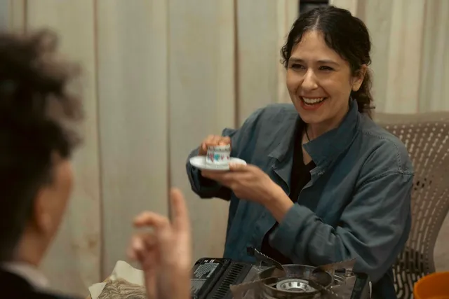 Tina Stefanou sits in a dark blue shirt with a big smile. She is inverting a cup on to a saucer about to read the coffee grounds fortune to someone who is sitting in front of her.