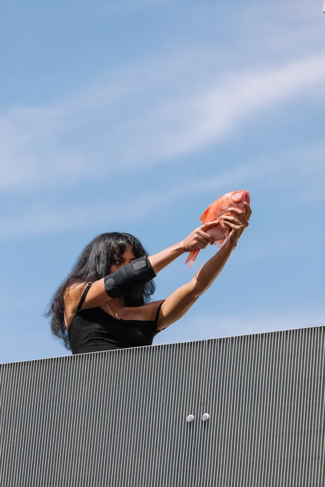 Ari posing with her arms out in front of her holding an orange fish like a gun.