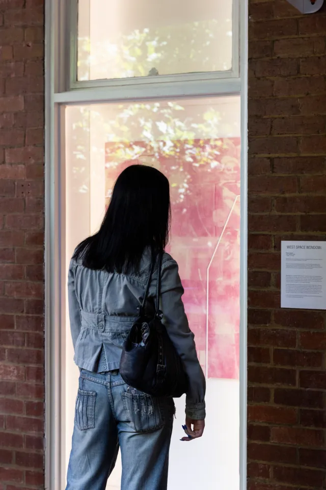 A woman in a shirt, pants and handbag stands in front of a large pink painting in the west space window