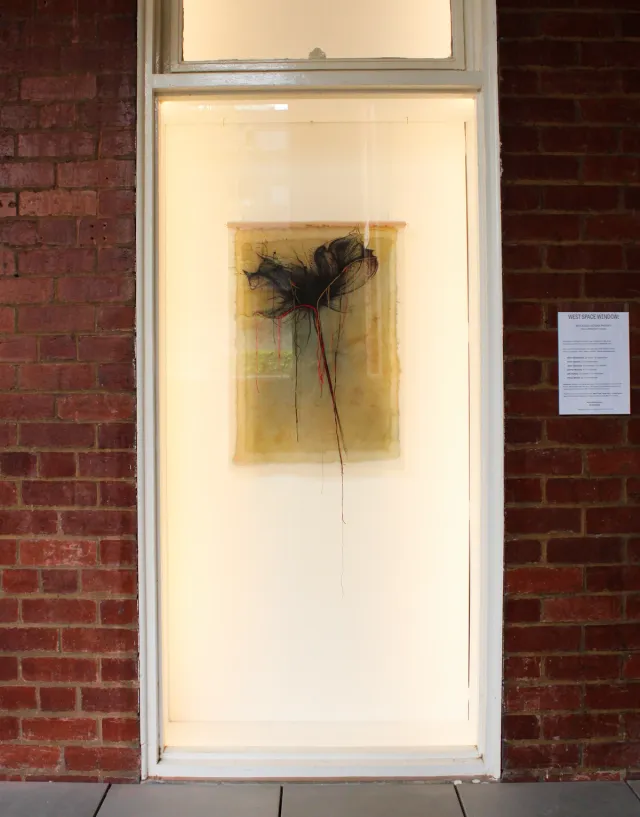 A flower made of black, red, and orange threads sewed on a yellow textile displayed in the West Space window, which is a recess in a red brick wall, with a label beside the window on the brick wall.
