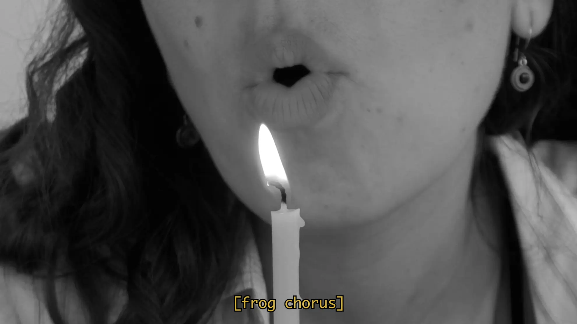 A close up film still of Tina with pursed lips blowing air on to a lit candle. A subtitle reads "frog chorus"