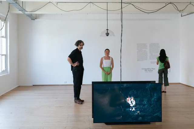 Two people watching video on the screen and one person reading texts on the wall at an exhibition.