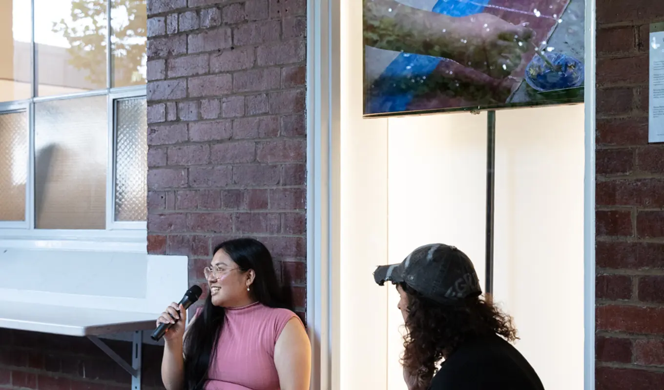 A person sitting on the floor talking to the microphone beside another person in front of an art display window.