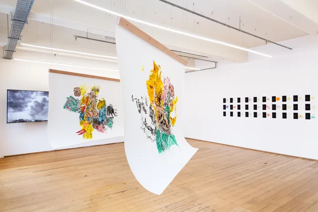 Two paintings hung from the ceiling, a TV and another installation in the back.