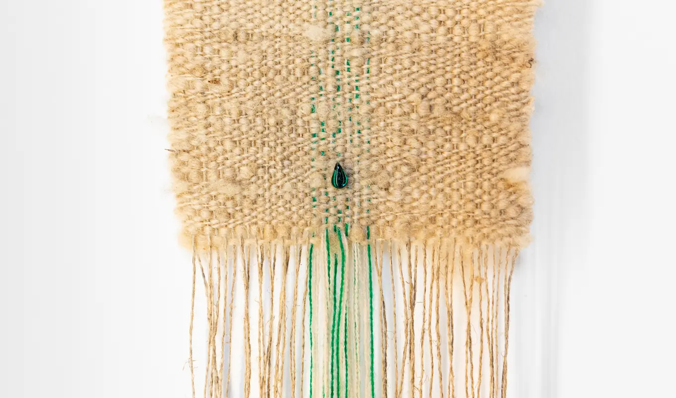 a beige coloured weaving with a few green threads and a single green gemstone towards the bottom of the weaving sits on a white wall. There are long threads coming out from the bottom of the weaving.