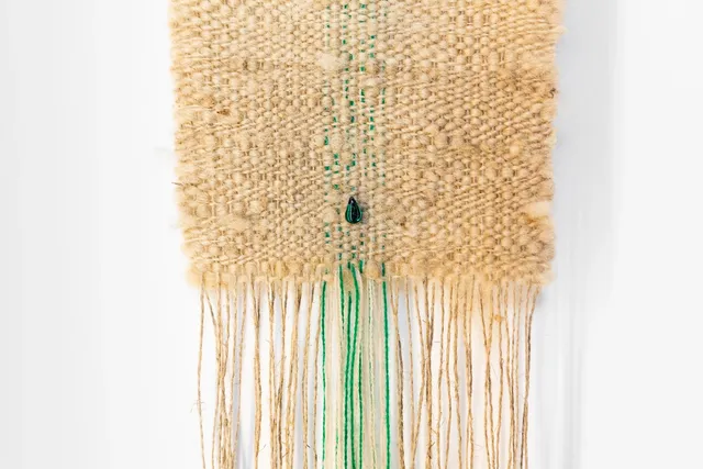 a beige coloured weaving with a few green threads and a single green gemstone towards the bottom of the weaving sits on a white wall. There are long threads coming out from the bottom of the weaving.
