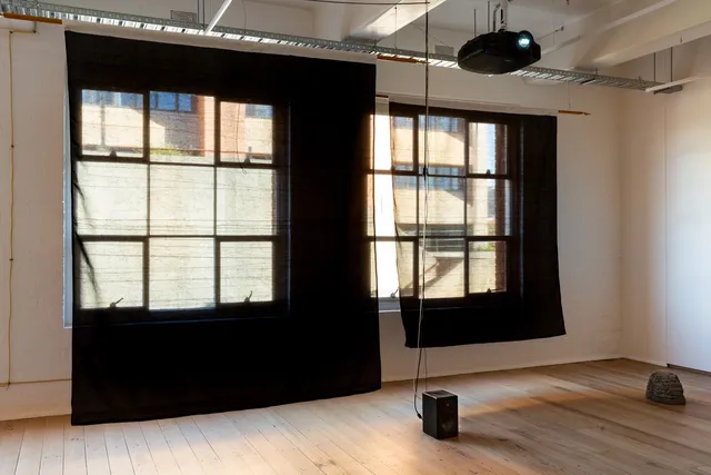 Installation view of "Big Pharmakon". The image shows two black semi-transparent sheets hanging from the ceiling in front of two large windows in West Space Gallery. In front of the sheets a small speaker and a plastic rock sit on the floor.