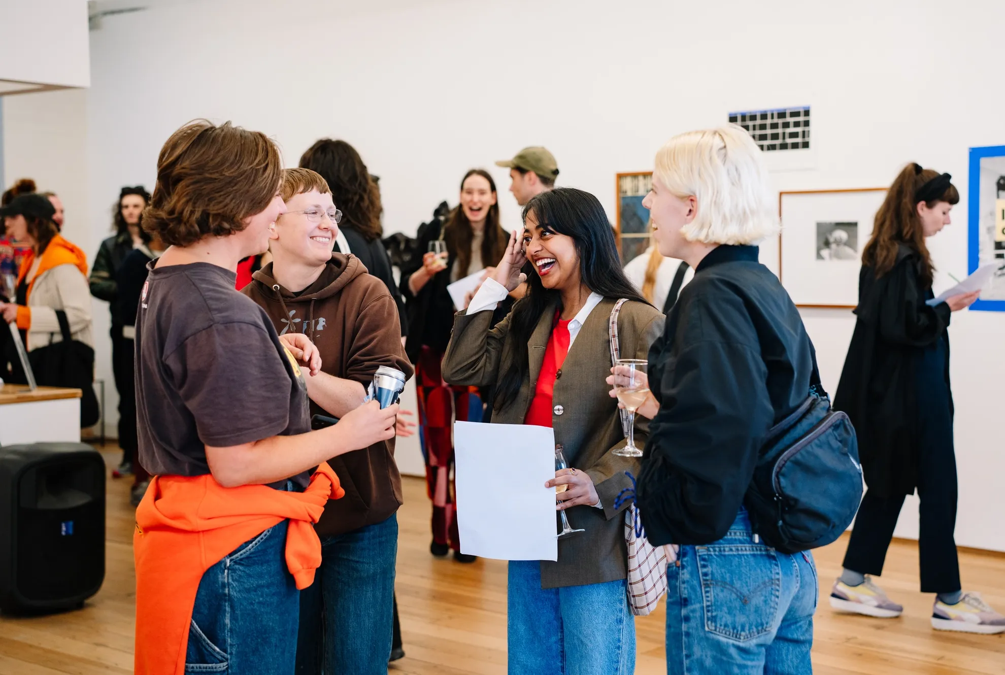 A group of 4 people foreground a larger crowd, all laughing in a crowded room. The walls are lined with two-dimensional artworks, all diverse.