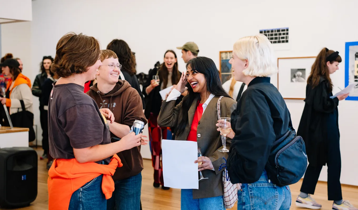 A group of 4 people foreground a larger crowd, all laughing in a crowded room. The walls are lined with two-dimensional artworks, all diverse.
