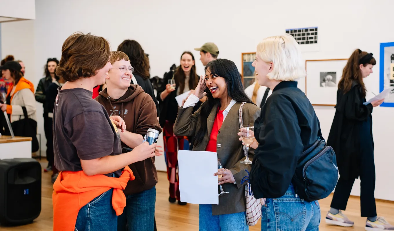 A group of 4 people foreground a larger crowd, all laughing in a crowded room. The walls are lined with two-dimensional artworks, all diverse.