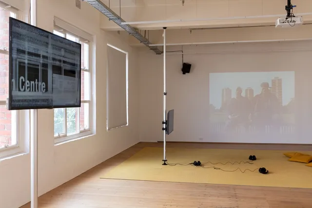Installation View of 13 Years showing 2 screens suspended away from the gallery wall , one has 3 pairs of headphones and some cushions in front of it. The back wall shows a projection of archival footage of 2 people sitting on a fence in front of 3 large apartment blocks.