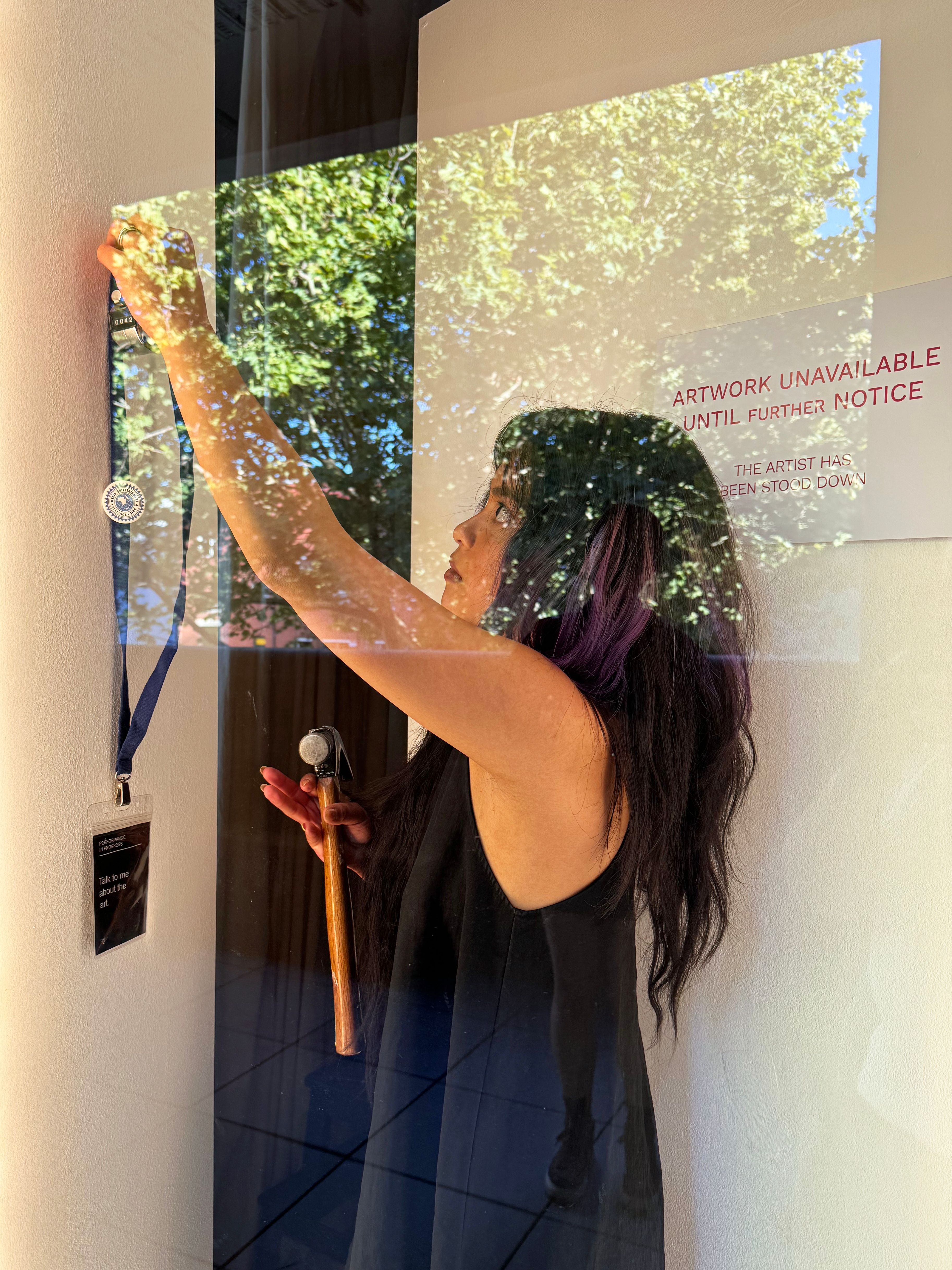 Bea Rubio-Gabriel behind a window holding a hammer in their right hand and a blue lanyard up to the wall in their left hand.