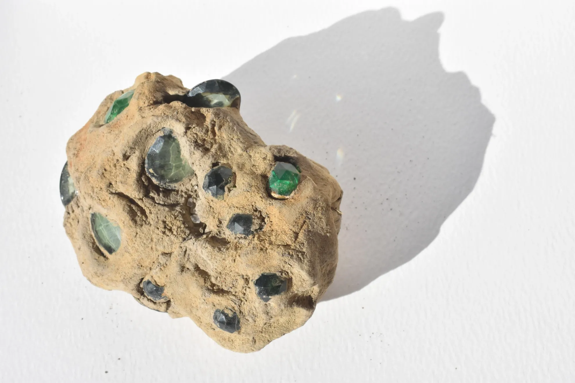 A small lump of unfired clay sits on a white background casting a shadow behind it. The lump of clay has been infused with an assortment of green plastic gemstones. The gemstones are sticking out in various areas of the clay.