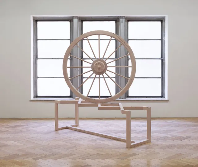 Front view of a large wooden sculpture or a wheel placed on a bench. The wheel is balancing on the intersection of two pieces of wood crossing over on the bench. The wheel is placed in front of a large window with natural light shinning through.
